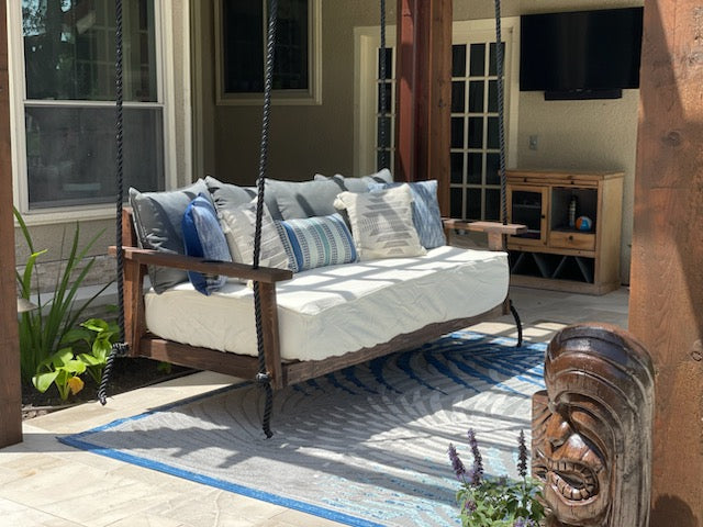 Wooden swing with cushions on a patio area with plants and a TV in the background. By Doorplace USA